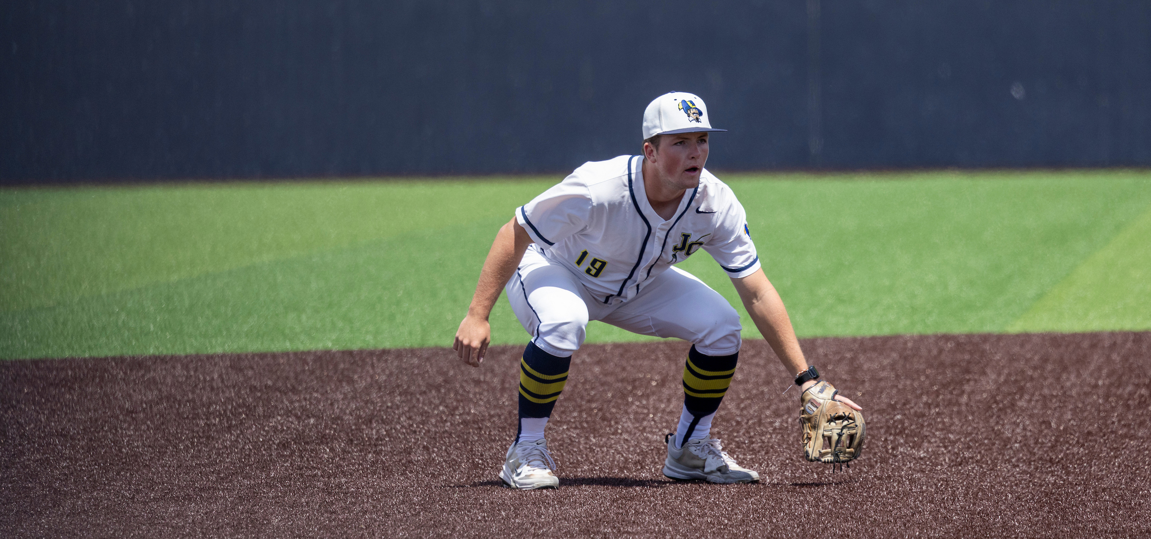Baseball player in a white uniform with the number 19 crouches in a ready fielding stance on a brown infield surface, wearing a glove low to the ground and facing forward. The player has a white cap with a logo, navy-and-gold striped socks, and white cleats. A green outfield and a dark fence form the background under bright daylight.