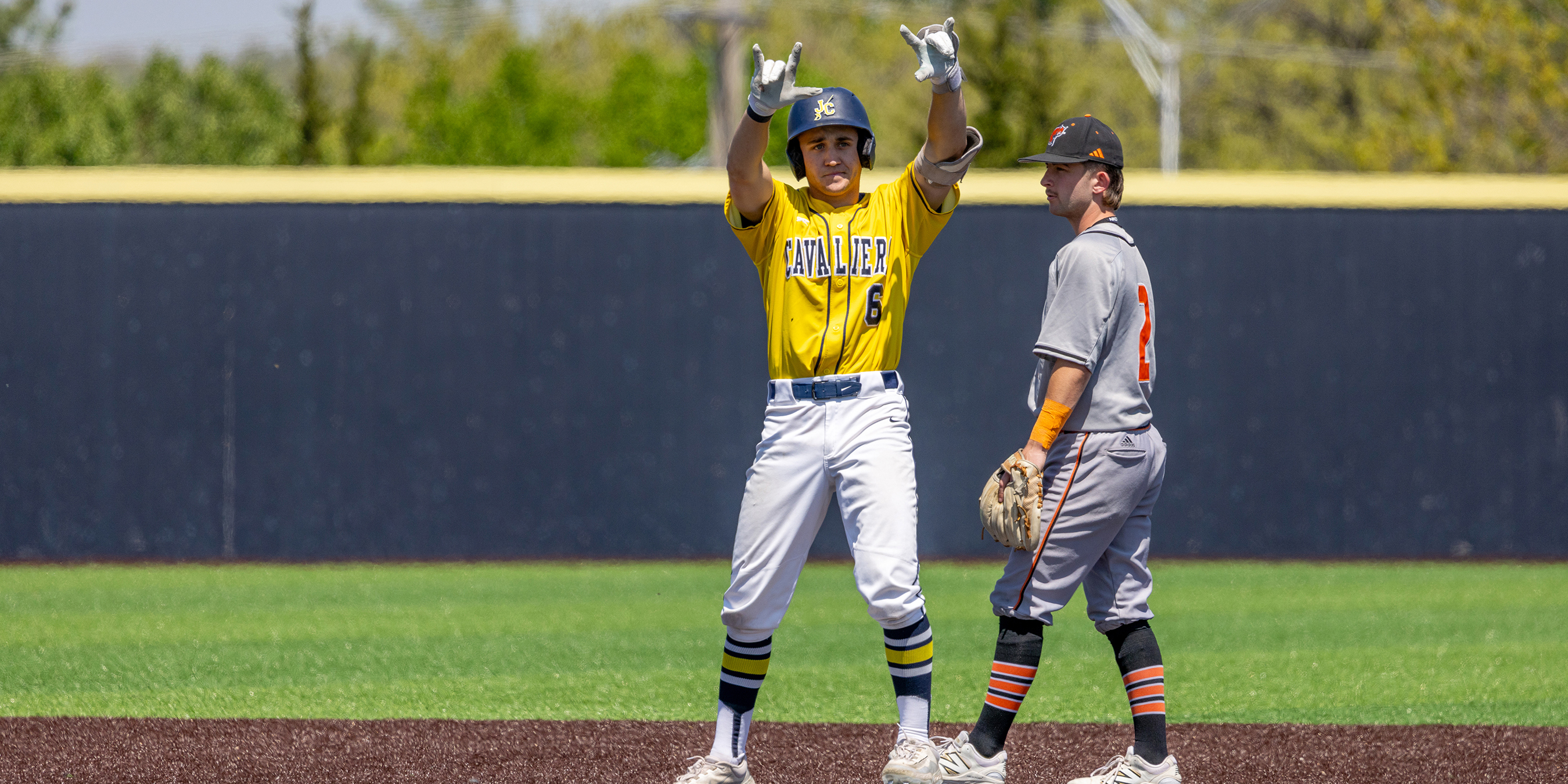In the photo, a baseball player, Ashton Hartwig, in a bright yellow jersey and white pants stands on a base, raising both arms in an enthusiastic celebratory gesture. His uniform features bold lettering across the chest, and he’s wearing matching striped socks and a helmet. Beside him, another player in a gray uniform with orange accents stands holding a glove, appearing to look toward the celebrating player. The scene takes place on a baseball field with green grass, a dark infield surface, and an outfield wall in the background under clear, sunny conditions.