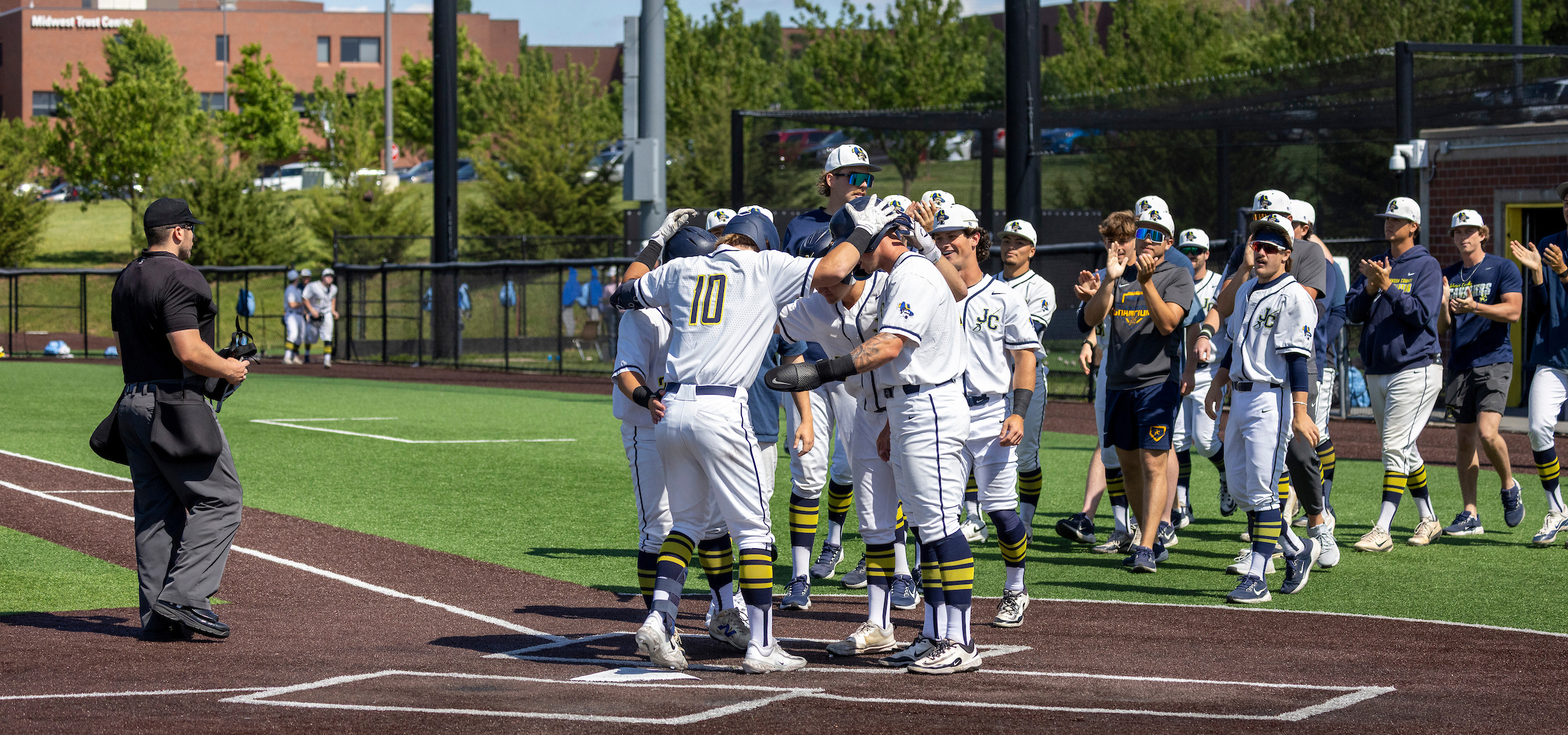 Baseball players in white uniforms with navy and yellow accents gather at home plate on a sunny day, tapping helmets in celebration after a run. An umpire dressed in black stands to the left near the foul line holding a face mask. Teammates in matching uniforms and casual athletic gear line up to the right along the fence, clapping. The field has green artificial turf and a dark brown base path, with a black chain-link fence and netting behind the plate. In the background, trees and a brick building labeled “Midwest Test Center” are visible under clear blue skies.