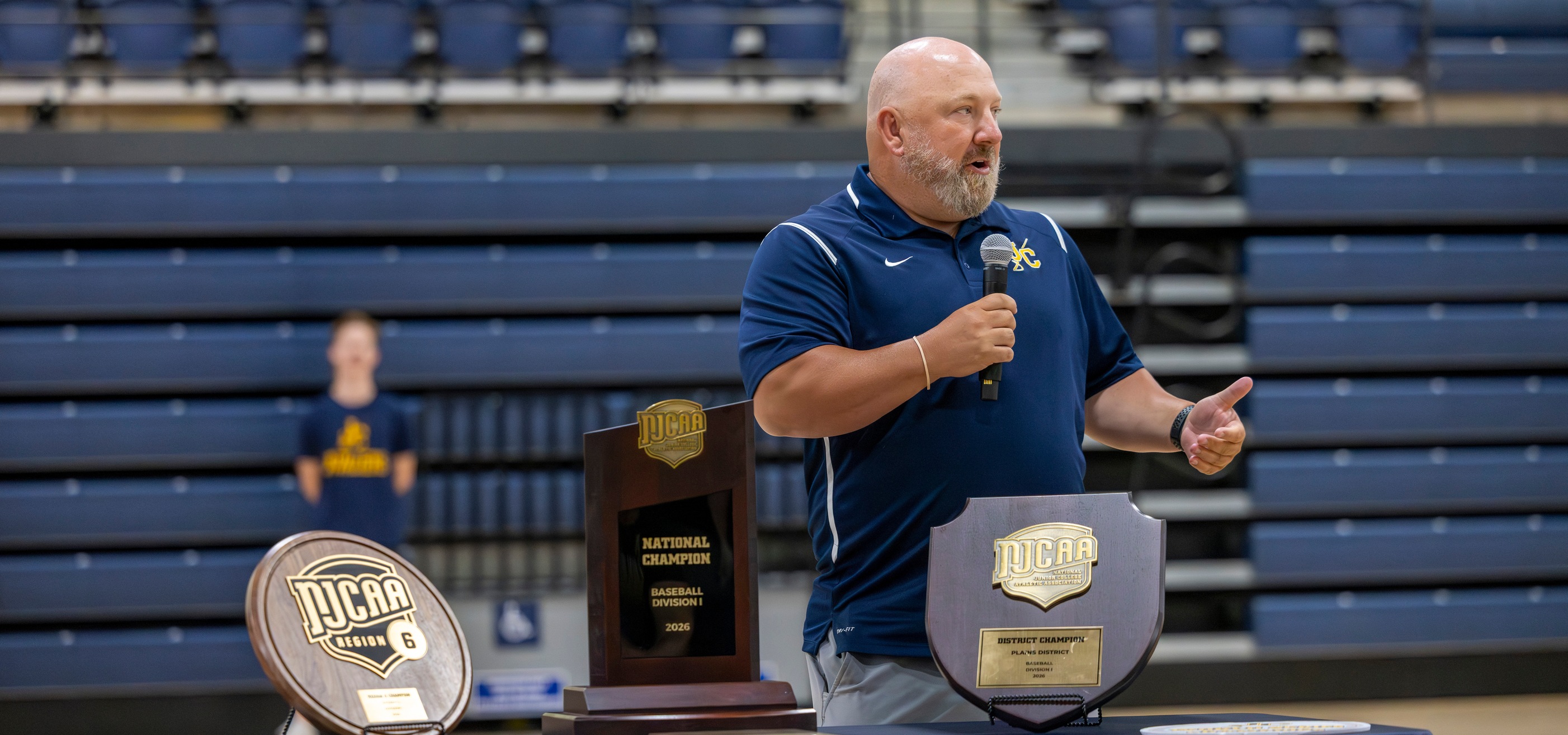 JCCC head coach Eric Horner stands in a gymnasium speaking into a handheld microphone while gesturing with one hand. The person wears a navy short-sleeve polo shirt with a small athletic logo and light-colored pants. In front of the speaker are three awards displayed on a table: a tall rectangular trophy with text reading “NJCAA National Champion Baseball Division I 2026,” a shield-shaped plaque labeled “NJCAA District Champion Plains District 2026,” and a round plaque marked “NJCAA Region 6.” Behind the speaker, rows of blue bleachers fill the background, and another individual appears blurred in the distance. The lighting is bright and even, typical of an indoor sports facility.