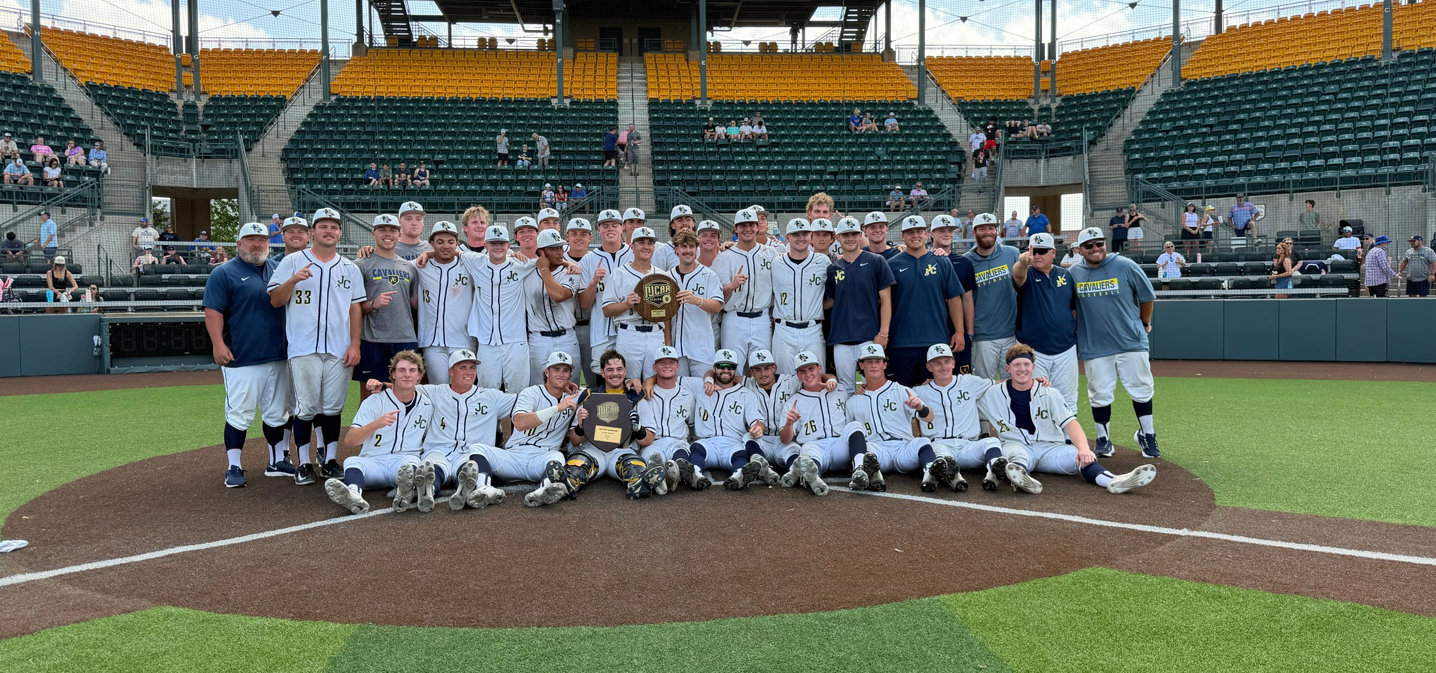 The Johnson County Community College baseball team poses for a group photo on the infield of a stadium. Around 30 players and several coaches wear white uniforms with dark lettering and caps, arranged in two rows with some standing and others sitting on the ground. Several players hold championship plaques, including one marked “NJCAA,” indicating a title win. The group is positioned near home plate on a brown infield surface with bright green turf around it. Behind the team, tiered stadium seating in green and yellow is partially filled with scattered spectators. A netted backstop and metal stadium structure frame the background under daylight conditions.
