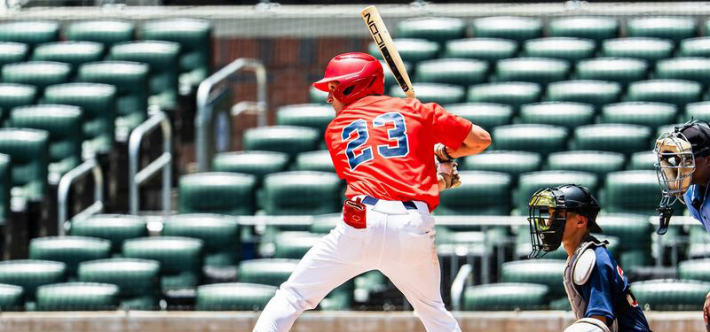 Baseball player wearing a red jersey with the number 23 and white pants stands in a batting stance at home plate, holding a bat over one shoulder. A catcher in dark protective gear crouches behind the batter, and part of an umpire is visible at the right edge of the frame. Rows of empty green stadium seats fill the blurred background on a bright day.