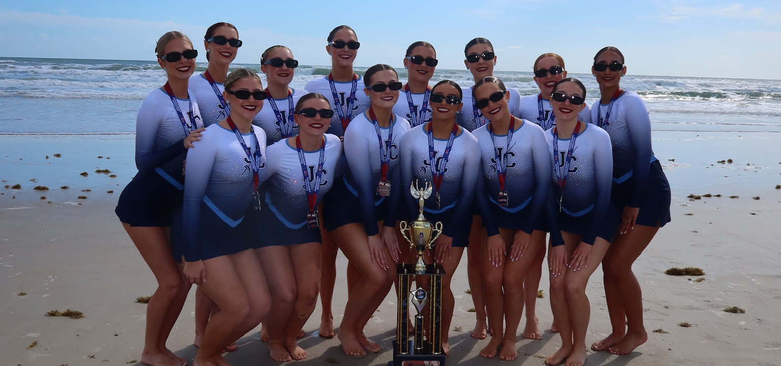 A group of about 15 people stands barefoot on a sandy beach in front of ocean waves, wearing matching long-sleeve performance uniforms and medals, gathered closely around a tall gold trophy placed in the center.