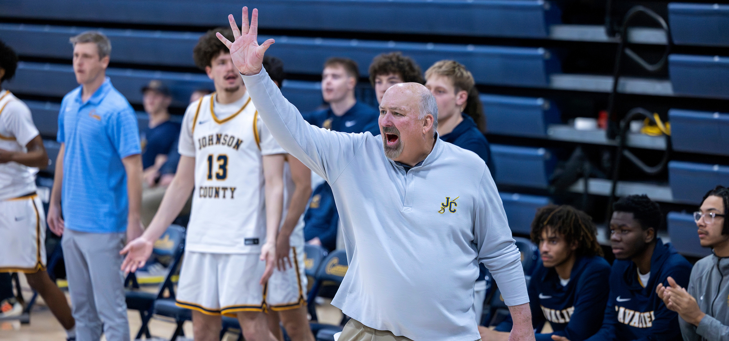 A basketball coach on the sidelines raises one hand with five fingers extended while calling out instructions during a game. The coach wears a light gray long-sleeve pullover with a “JC” logo and stands in front of a team bench. Behind the coach, players in white uniforms with “Johnson County” and the number 13 visible react and gesture, while others in navy warmups sit and watch. Blue bleachers fill the background of the indoor gymnasium.