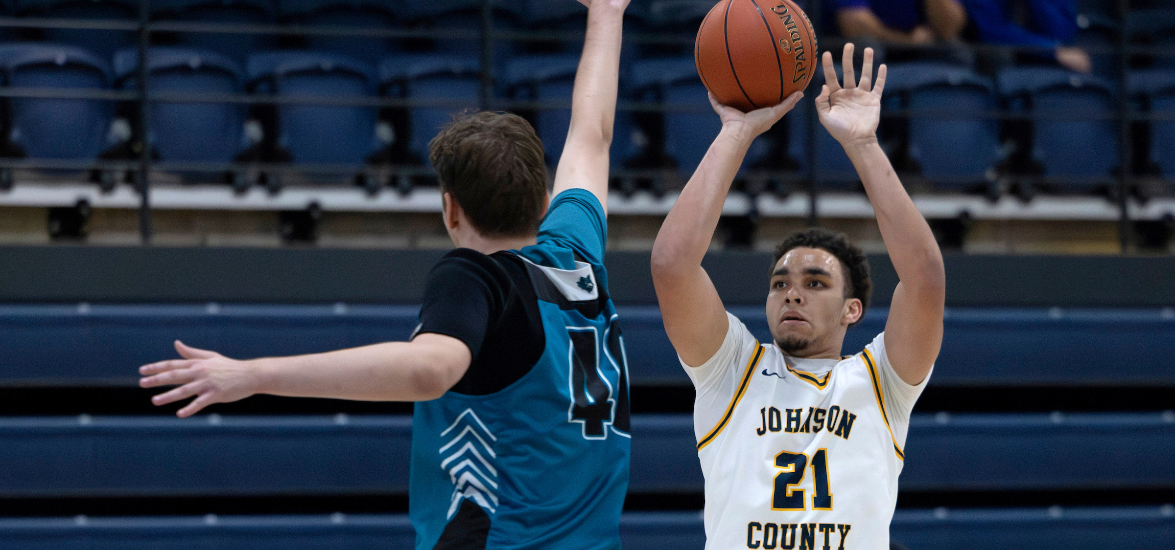 The photo shows an indoor basketball game in progress. One player wearing a white jersey with yellow trim and the text “JOHNSON COUNTY” along with the number 21 is in the act of shooting the basketball. The player is holding the ball above their head with both hands, preparing for a shot. Another player in a teal and black jersey with the number 14 is positioned defensively, with one arm extended upward to contest the shot. The background features rows of blue stadium seats and a railing, indicating the game is taking place in a gymnasium or sports arena. The lighting is bright and evenly distributed, typical of an indoor sports facility.