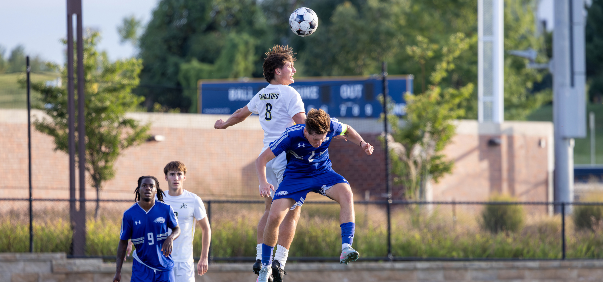 The photo captures an intense moment during a soccer match. Two players are airborne, competing for a header. The player in white, wearing jersey number 8 with the word “CAVALIERS” visible on the back, is slightly higher than the player in blue, who is also jumping to challenge for the ball. The soccer ball is in mid-air above them.
In the background, there are two additional players in blue jerseys standing on the field, and a scoreboard partially visible behind a brick wall. The setting appears to be an outdoor soccer field with green trees and a clear sky, suggesting good weather conditions. The overall scene conveys athletic action and competition.
