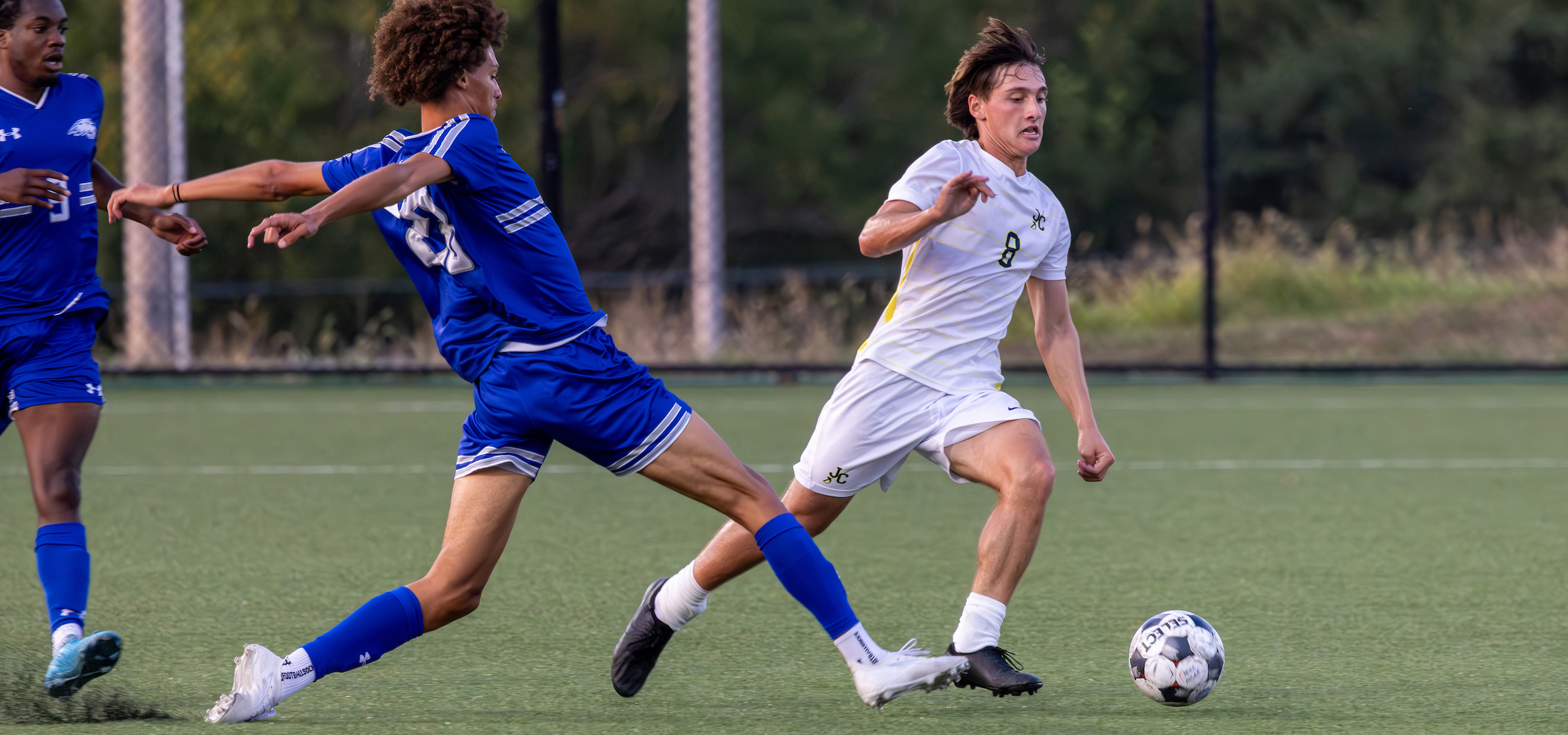 The photo shows an intense soccer match on an outdoor field with artificial turf. Two players are the main focus:

Player in blue: Wearing a blue jersey and shorts with white trim, along with blue socks and white cleats. This player is in a defensive stance, extending one leg in an attempt to block or tackle the ball.
Player in white: Wearing a white jersey with yellow side accents and white shorts, along with white socks and black cleats. The jersey has the number 8 visible. This player is controlling the ball, which is a standard black-and-white patterned soccer ball, and appears to be moving forward aggressively.

In the background, there is a blurred view of green foliage and a black fence, suggesting the game is taking place on a fenced-in soccer field. The lighting indicates it might be late afternoon or early evening, with natural light illuminating the scene.
