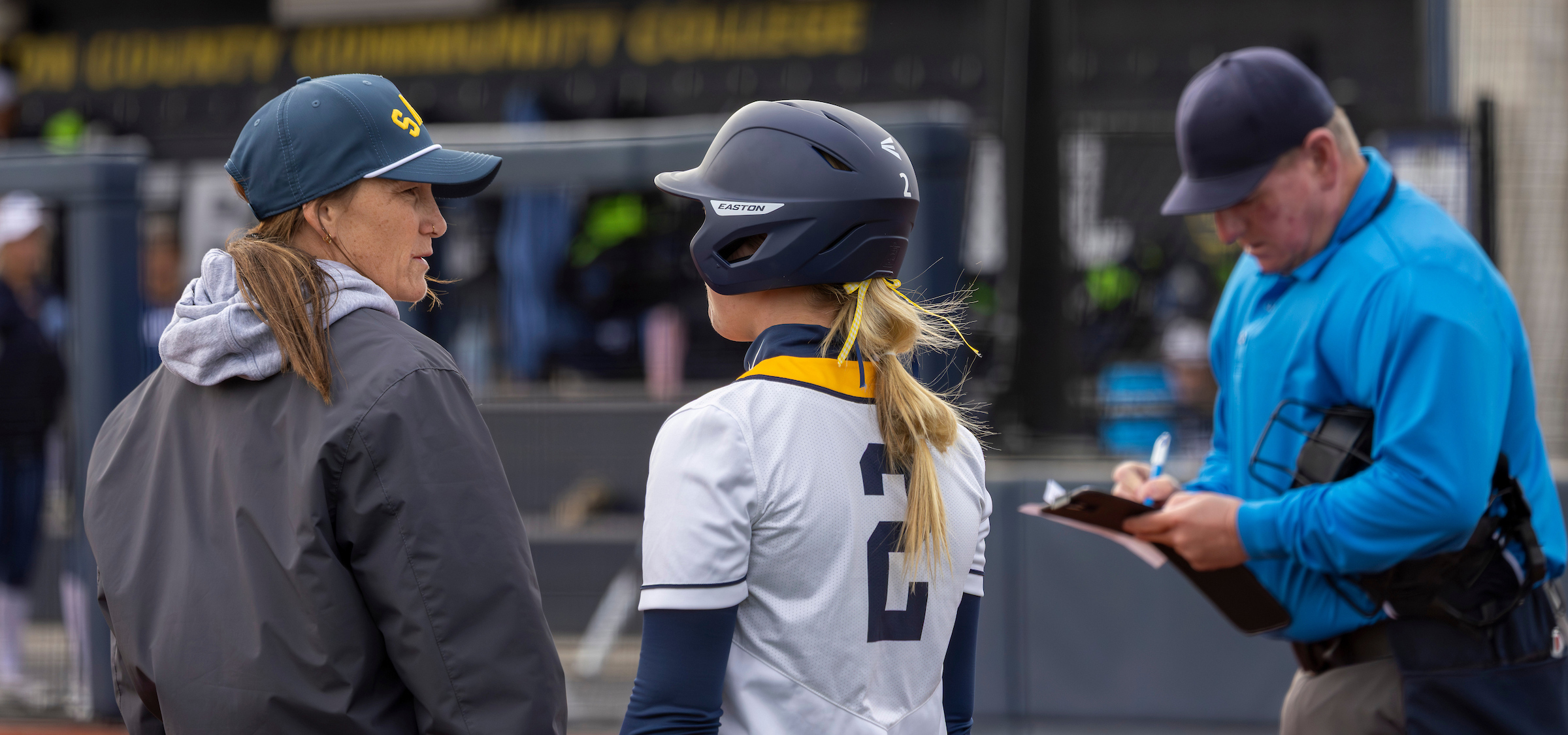 The image shows three individuals standing near a softball field. One appears to be a player wearing a navy helmet, a white uniform top with dark sleeves, and the number 2 on the back. This player is facing two adults—one wearing a dark jacket and a ball cap, and the other dressed in a bright blue umpire-style shirt and cap.
The player seems to be engaged in conversation with the person in the dark jacket, who is turned slightly toward the player. The individual in the blue shirt is holding a clipboard and appears to be writing something down.
In the background, parts of the softball complex are visible, including fencing, dugout areas, and signage with yellow lettering. The scene looks like it is taking place just outside the playing field, possibly during a discussion before or after an at?bat or between innings.