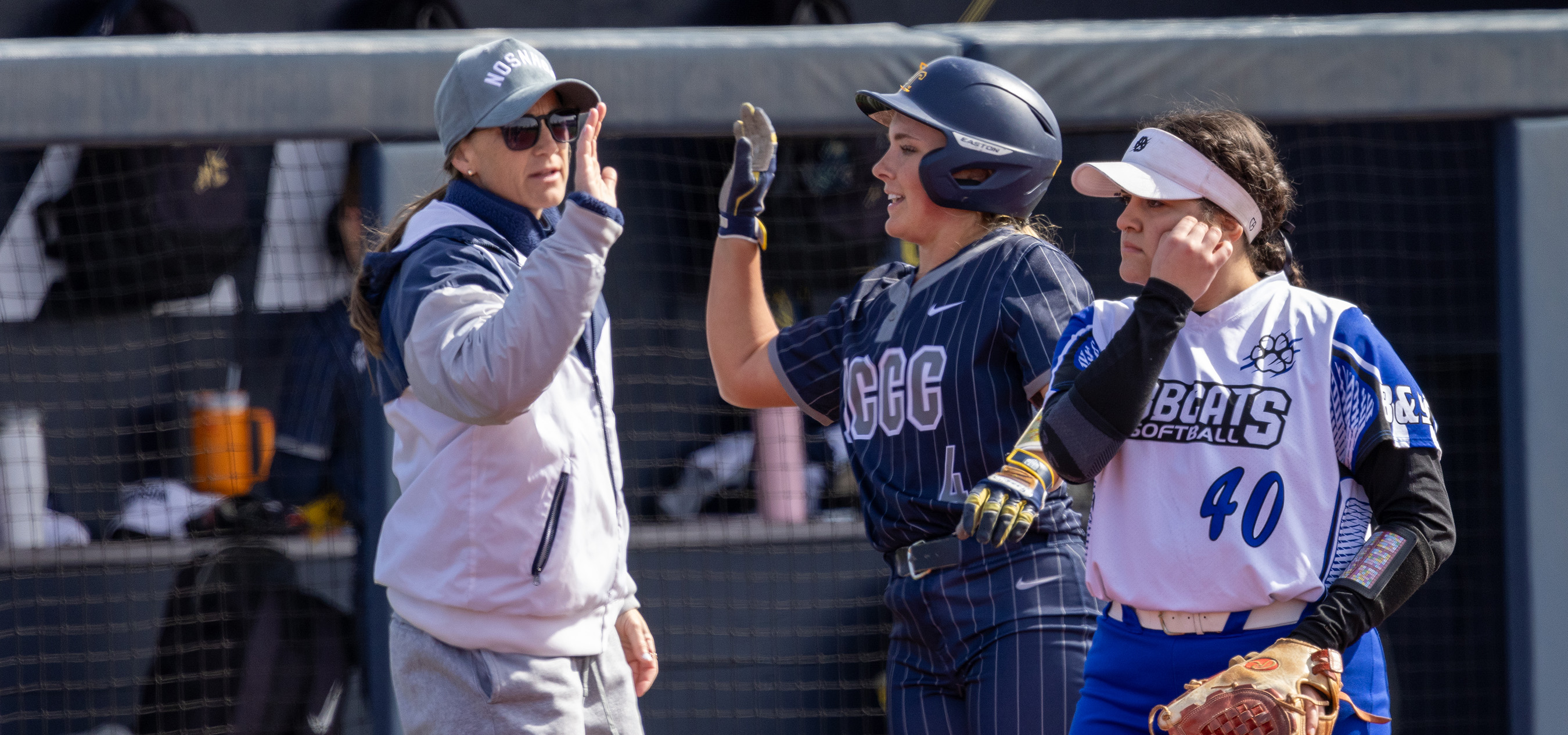 Softball scene at a field: a player in a dark, pinstriped uniform with the number “4” and a batting helmet raises a gloved hand to high-five a coach in a gray jacket, cap, and sunglasses near the dugout fence. Another player in a white-and-blue “BEARCATS SOFTBALL” jersey with the number “40” stands beside them holding a glove. Nets, equipment, and teammates appear in the background under bright daylight.
