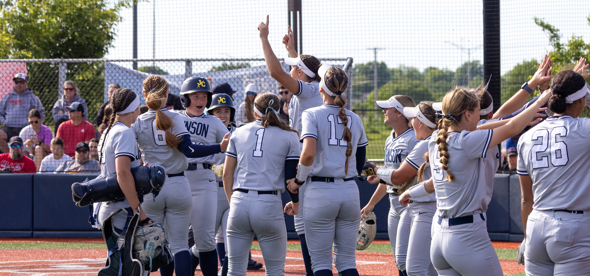 Softball team in gray uniforms gathers in a circle on a turf field near home plate, raising hands and high-fiving around a helmeted player wearing a “JC” logo. A catcher stands at left holding gear, while other players with visible numbers (including 1, 12, 5, and 26) face inward. Spectators sit behind a chain-link backstop fence in the background under bright daylight.