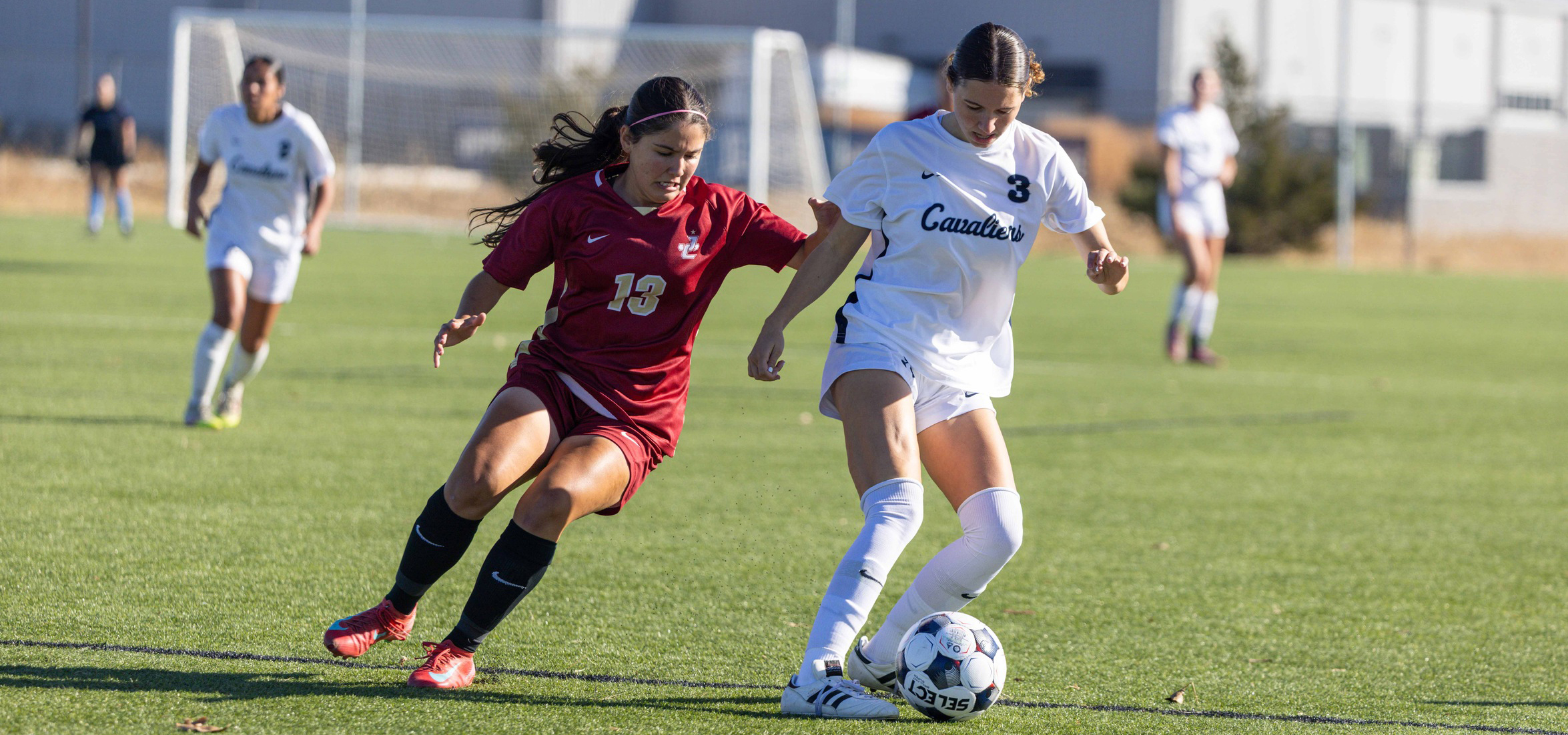 The photo shows an outdoor soccer match on a well-maintained grass field. Two players are in the foreground actively competing for possession of the ball.

The player on the right is wearing a white uniform with the word “Cavaliers” printed on the front and the number 3 visible. This player is controlling the ball with the right foot and appears to be moving forward.
The player on the left is wearing a maroon uniform with the number 13 displayed prominently. This player is positioned slightly behind and to the side, appearing to challenge for the ball.
Both players are wearing cleats and shin guards, and the soccer ball is marked with visible text and patterns.
In the background, there is a goalpost with a net, additional players in white uniforms, and a blurred building structure beyond the field. The lighting suggests a sunny day with clear skies.