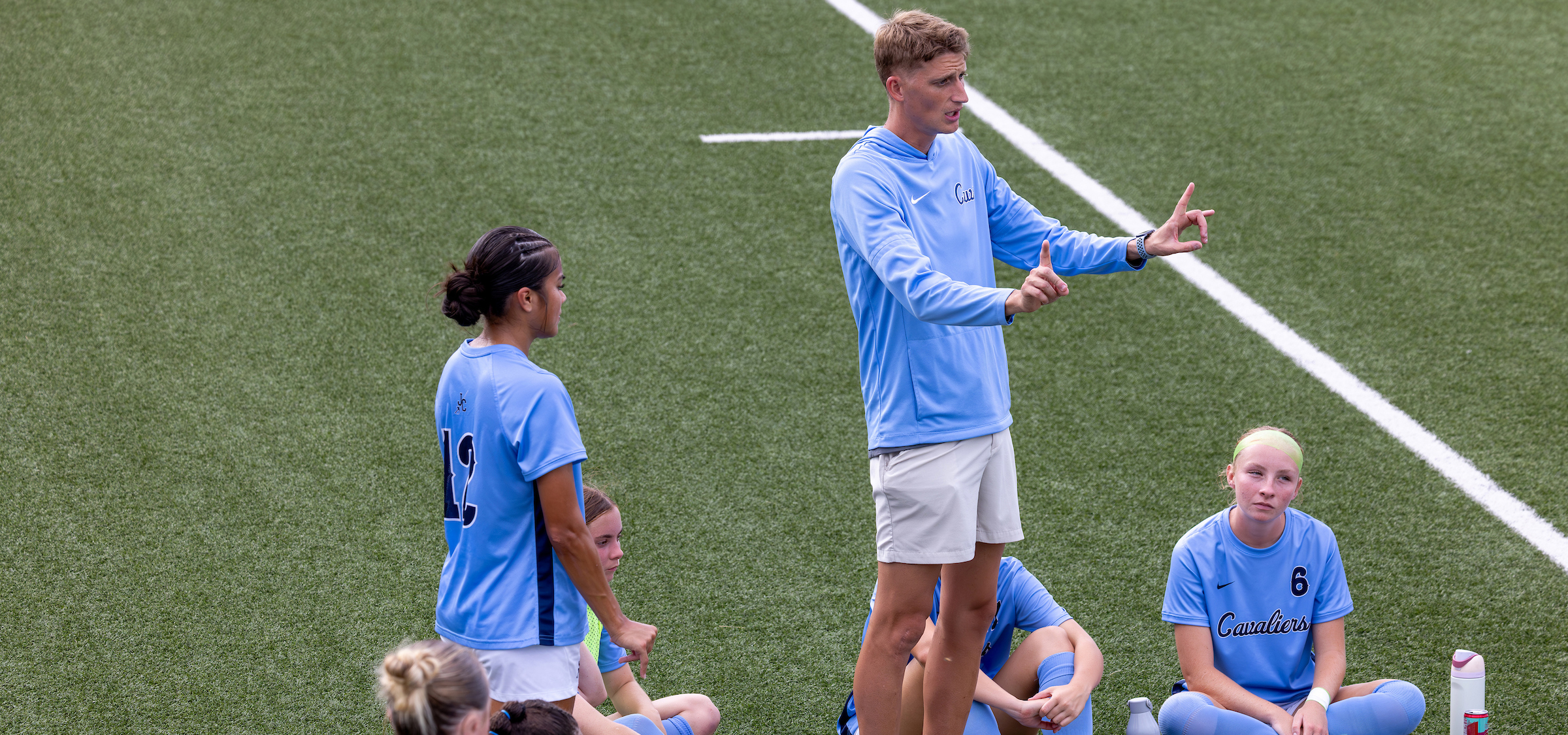 The image shows a scene on a sports field with artificial turf. A group of individuals wearing light blue athletic uniforms is gathered near the sideline. One individual is standing and appears to be addressing the group, using hand gestures that suggest giving instructions or explaining something. This individual is wearing a long?sleeve light blue top and light-colored shorts.
Several others are seated or kneeling on the turf nearby, wearing similar light blue jerseys with dark numbers visible on the backs or fronts. One jersey shows the number 19, and another shows 6. A white water bottle with red accents is placed on the ground next to one of the seated individuals.
The overall setting suggests a team meeting or timeout during a sports practice or game. The background consists entirely of the artificial turf field, with a white boundary line running diagonally through the image.