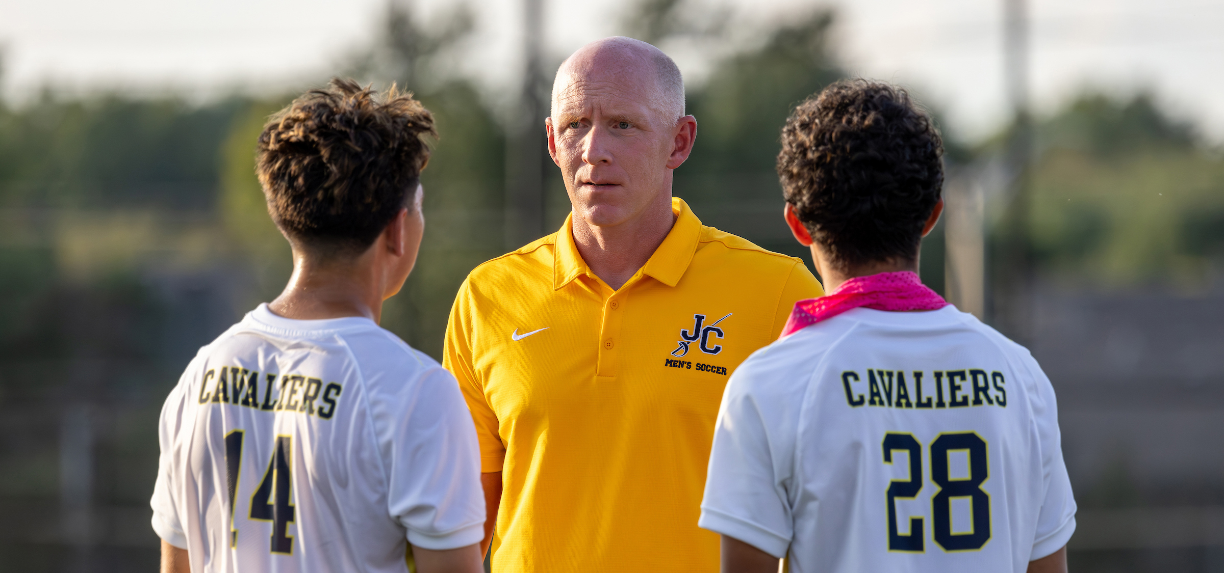 The photo shows soccer coach Jason Wichman standing between two players on what appears to be an outdoor soccer field. The coach is facing the camera and wearing a bright yellow polo shirt with a logo that reads “JC Men’s Soccer.” His expression is focused and serious, suggesting he is giving instructions or having a conversation with the players.
On either side of him are two players seen from behind, both wearing white soccer jerseys with “Cavaliers” printed across the upper back. One player wears number 14, and the other wears number 28. The player on the right also has a pink bandana or neck gaiter around his neck.
The background is softly blurred, showing trees and goal structures, indicating the photo was taken during practice or a match in daylight. The overall mood suggests a coaching moment, possibly a discussion or strategy talk during a break in play.