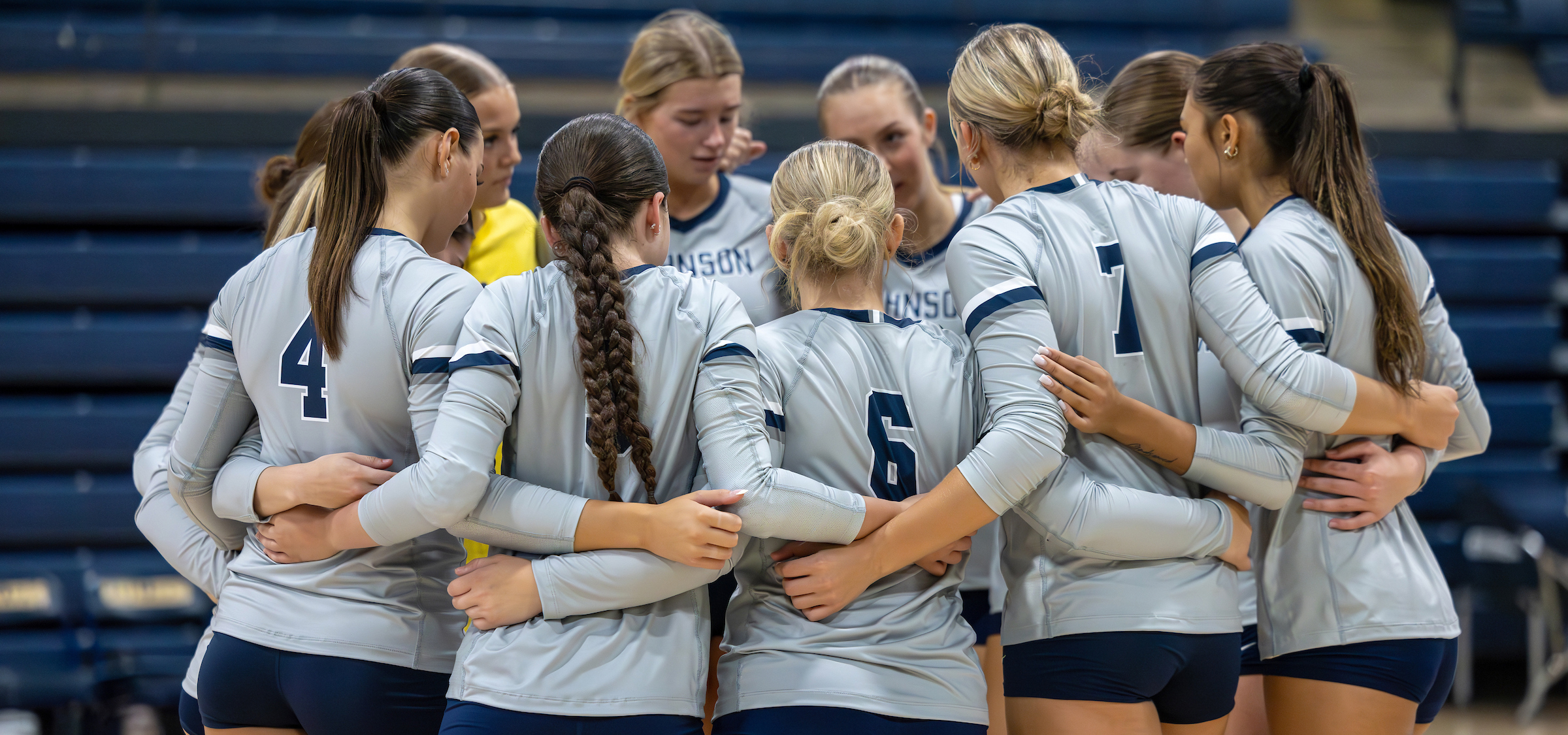 The photo depicts a group of volleyball players in a team huddle. They’re wearing coordinated uniforms—long-sleeved jerseys and shorts—with visible jersey numbers like 4, 6, and 7. The players have their arms around each other, indicating a moment of unity or strategizing before or during a game. The setting is an indoor sports facility, with blue bleachers visible in the background.